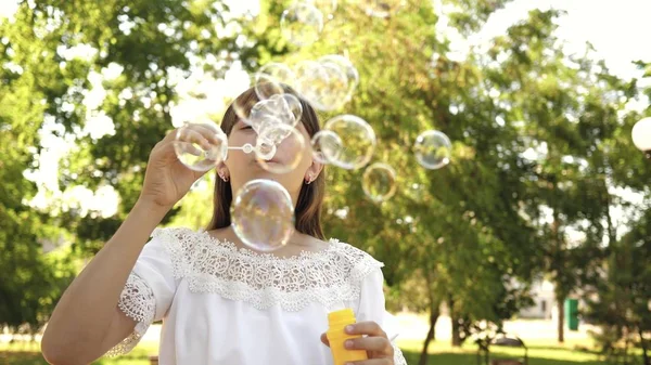 Ung flicka leker i parken och blåser bubblor i kameralinsen. Slow motion. Vacker flicka blåser såpbubblor i parken i vår, sommar och leende. — Stockfoto