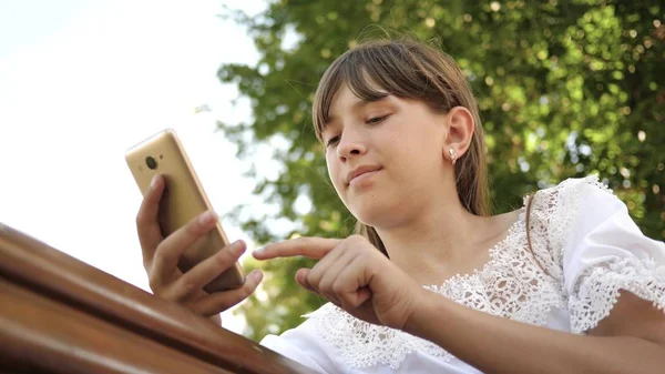 快乐的女孩使用智能手机翻转网页在互联网上的公园在长凳上。特写。在植物园的年轻的千年妇女, 在手机上做手势显示. — 图库照片