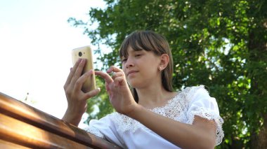 Bir akıllı telefon kullanan genç bir kız güzel bir yeşil parkta bir bankta bir mektup yazıyor. Arboretum 'da genç Milenyum kadın, telefon ekranında jestler yapma.