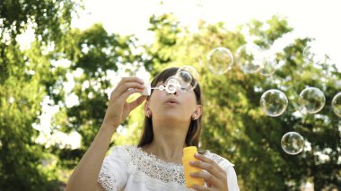 Genç Kız parkta oynarken ve kabarcıklar objektife içine üfleme. Ağır çekim. İlkbahar, yaz ve gülümseyen içinde parkta sabun köpüğü üfleme güzel kız.