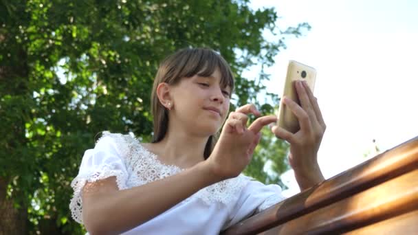 Ung tusenåriga kvinna i Arboretum, göra gester på telefonens display. En ung flicka med en smartphone är att skriva ett brev på en bänk i en vacker grön park. — Stockvideo