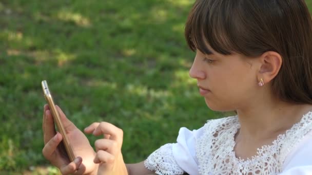 Hermosa chica usando un teléfono inteligente está escribiendo una carta en un banco en un hermoso parque verde. En cámara lenta. Joven mujer milenaria en el Arboreto, haciendo gestos en la pantalla del teléfono . — Vídeo de stock