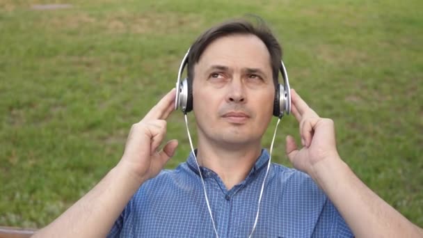 Joven guapo escuchando música desde su teléfono inteligente con auriculares, bailando afuera en el parque en un banco en el centro de Europa — Vídeo de stock