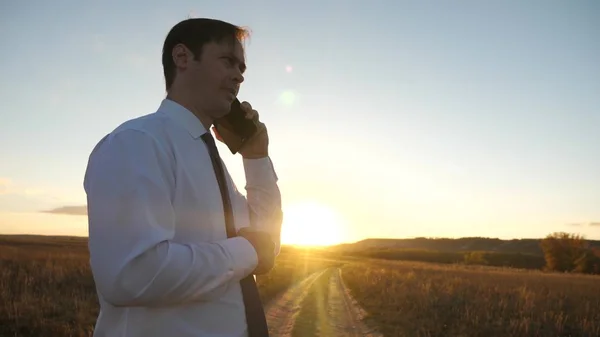 Homem de negócios falando em um smartphone contra o céu. homem de gravata com um tablet no parque ao pôr-do-sol. mensagem móvel falando no telefone inteligente. empresário em uma estrada de campo com um telefone celular — Fotografia de Stock