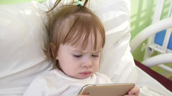 可爱的小女孩躺在儿童医院的床上, 在智能手机上看有趣的动画片. — 图库照片