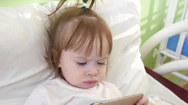 可爱的小女孩躺在儿童医院的床上, 在智能手机上看有趣的动画片. — 图库照片