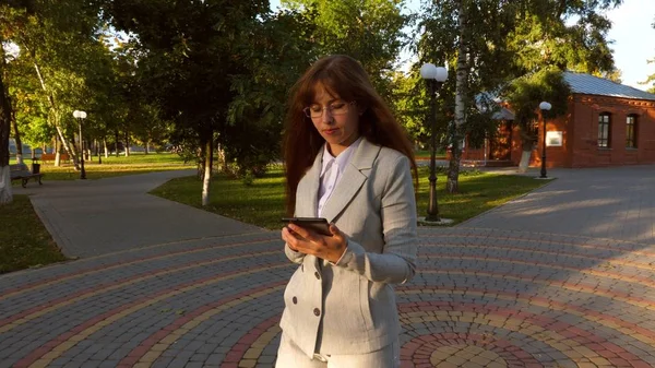 Bela mulher de negócios com óculos andando ao longo do beco no parque com tablet em suas mãos, menina em um terno de negócios leve vai trabalhar . — Fotografia de Stock