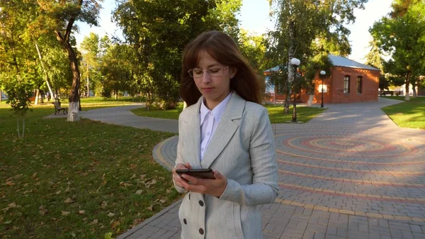Hermosa mujer de negocios con gafas caminando por el callejón en el parque con la tableta en sus manos, chica en un traje de negocios ligero va a trabajar . —  Fotos de Stock