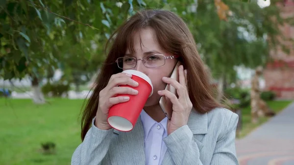 Hermosa chica está bebiendo café y hablando en el teléfono inteligente, chica se ríe. joven mujer en gafas en traje de luz va en el parque en verano — Foto de Stock