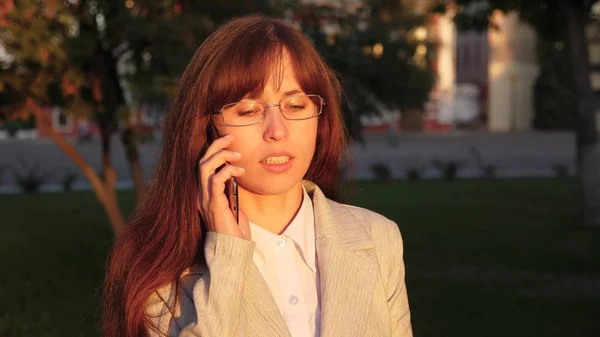 Mujer de negocios con gafas en traje de luz va a trabajar. mujer camina por una calle de la ciudad iluminada por rayos de sol y habla en un teléfono inteligente . —  Fotos de Stock