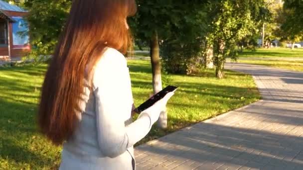 Meisje met tablet in een lichte pak gaat aan het werk. mooie zakenvrouw wandelingen langs steegje in park met tablet in haar handen. — Stockvideo