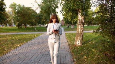 güzel iş kadını sokakta park tablet ile birlikte onun elinde yürür, hafif iş takım elbiseli kız işe gider.