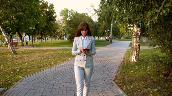 Hermosa mujer de negocios camina por el callejón en el parque con la tableta en sus manos, chica en un traje de negocios ligero va a trabajar . —  Fotos de Stock