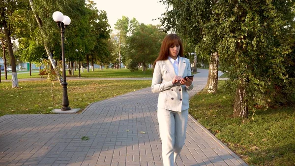 Hermosa mujer de negocios camina por el callejón en el parque con la tableta en sus manos, chica en un traje de negocios ligero va a trabajar . —  Fotos de Stock