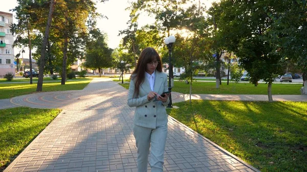 Chica sensual en un traje de negocios ligero va a trabajar. hermosa mujer de negocios caminando por el callejón en el parque con una tableta en sus manos . —  Fotos de Stock