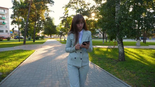 Chica sensual en un traje de negocios ligero va a trabajar. hermosa mujer de negocios caminando por el callejón en el parque con una tableta en sus manos . —  Fotos de Stock