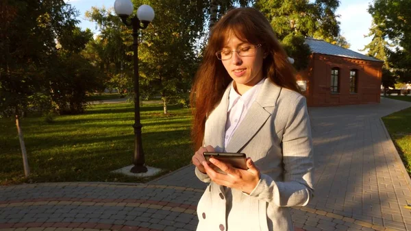 Krásná obchodnice s brýlemi pěší uličkou v parku s tabletem v ruce, dívka v lehké odhalenou jde do práce. — Stock fotografie