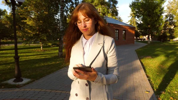 Schöne Geschäftsfrau mit Brille, die mit Tablet in der Hand die Gasse im Park entlanggeht, Mädchen im leichten Businessanzug zur Arbeit. — Stockfoto