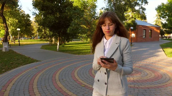 Hermosa mujer de negocios con gafas caminando por el callejón en el parque con la tableta en sus manos, chica en un traje de negocios ligero va a trabajar . —  Fotos de Stock