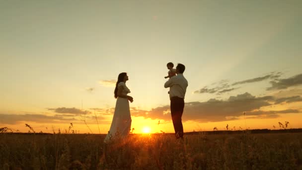 Papá tiene en sus manos a una pequeña y feliz hija y se la da a su madre. hija juega con mamá y papá al sol. concepto de familia feliz y la infancia. familia feliz descansando en el parque en los rayos de la puesta del sol — Vídeos de Stock