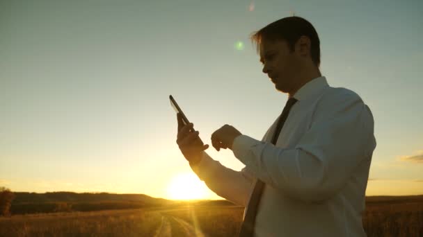 Людина в краватці з планшетом і перевірка електронної пошти в парку на заході сонця. бізнесмен, що працює з планшетом, смартфон проти неба. мобільне повідомлення на екрані смартфона. бізнесмен на заміській дорозі з навігатором — стокове відео
