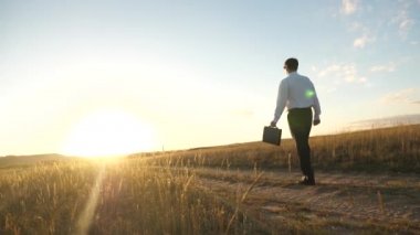 Girişimci bir kırsal bölgede çalışıyor. Çiftçi kara gün batımında olup olmadığını denetler. Güneş gözlüğü yavaş motion.businessman elinde bir çanta ile ülke yolda gidiyor. tarıma dayalı işletme kavramı