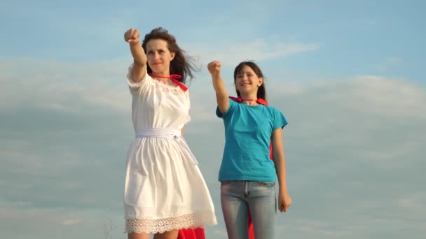 Superhéroes del trabajo en equipo. dos chicas con capas rojas de superhéroes se paran contra un cielo azul, el viento infla una capa. Mamá y su hija juegan a los superhéroes. concepto familiar feliz . — Vídeo de stock