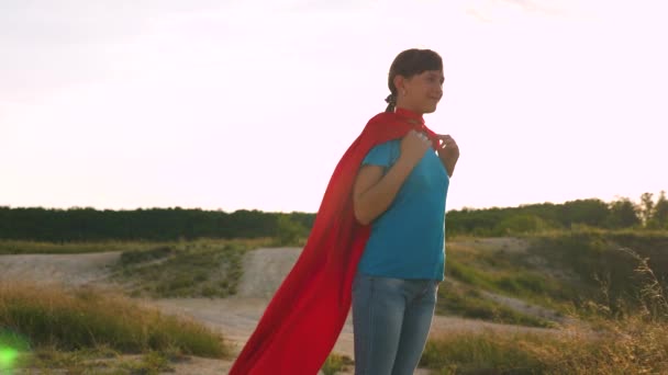 Hermosa chica superhéroe de pie en el campo en una capa roja, capa revoloteando en el viento. En cámara lenta. Una joven sueña con convertirse en un superhéroe. chica camina en una expresión de capa roja de sueños — Vídeo de stock