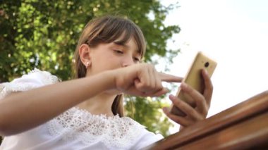 bir parkta bir bankta Internet sayfaları aracılığıyla saygısız smartphone kullanan mutlu kız. yakın çekim. Bin yıllık kadında genç jestleri ekranın üzerinde yapma Botanik Bahçesi,.