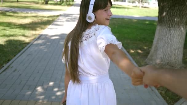 Teamwerk van een verliefde paar. meisje en jongen reizen naar muziek hand in hand. Slow-motion. Het meisje in hoofdtelefoons glimlacht en leidt de geliefde man bij de hand in het park een lentedag. — Stockvideo