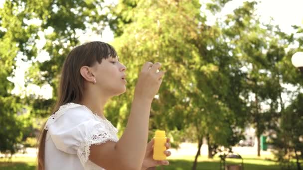 Fröhliches Mädchen, das im Frühling, Sommer und lächelnd schöne Seifenblasen im Park pustet. Zeitlupe. junges Mädchen unterwegs durch die Stadt im Park. — Stockvideo