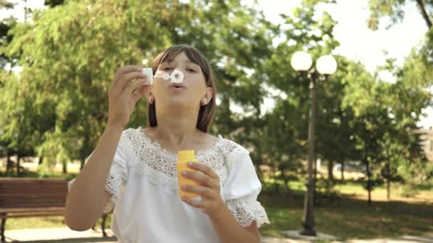 Mladá dívka hraje v parku a vyfukuje bubliny do objektivu fotoaparátu. Zpomalený pohyb. Krásná dívka foukání mýdlové bubliny v parku na jaře, v létě a s úsměvem. — Stock video