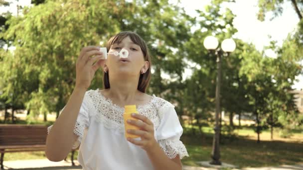 Niña jugando en el parque y soplando burbujas en la lente de la cámara. En cámara lenta. Hermosa chica soplando burbujas de jabón en el parque en primavera, verano y sonriendo . — Vídeo de stock