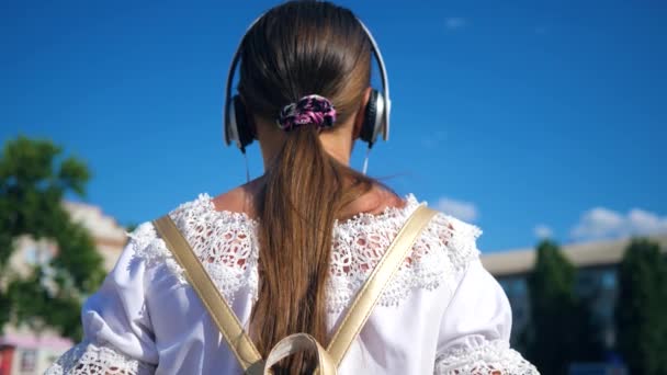 Menina adolescente em vestido branco com cabelos longos viaja pela cidade contra o céu azul. Movimento lento. Uma jovem caminha pela rua da cidade com fones de ouvido e ouve música . — Vídeo de Stock