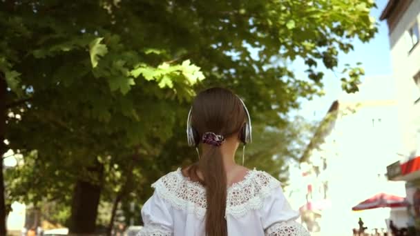 Chica en un vestido blanco con el pelo largo viaja por la ciudad. En cámara lenta. chica feliz caminando por la calle de la ciudad en auriculares y escuchando música. adolescente salida en la ciudad . — Vídeo de stock