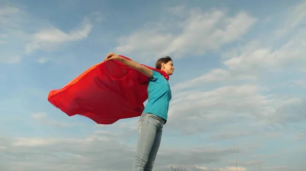 Mooi meisje superhero staande op het veld in een rode mantel, mantel wapperen in de wind. Slow-motion. meisje droomt ervan om een superheld. jong meisje permanent in de expressie van een rode mantel van dromen — Stockfoto