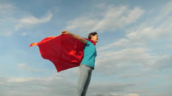 Mooi meisje superhero staande op het veld in een rode mantel, mantel wapperen in de wind. Slow-motion. meisje droomt ervan om een superheld. jong meisje permanent in de expressie van een rode mantel van dromen — Stockfoto