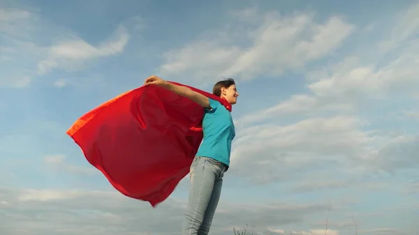 Bela menina super-herói em pé no campo em um manto vermelho, vacilante manto no vento. Movimento lento. menina sonha em se tornar um super-herói. jovem de pé em um manto vermelho expressão de sonhos — Fotografia de Stock