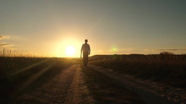 işadamı güneş gözlüğü elinde bir çanta ile ülke yolda gidiyor. Girişimci bir kırsal bölgede çalışıyor. bir çiftçi kara gün batımında olup olmadığını denetler. tarıma dayalı işletme kavramı.