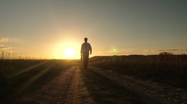 işadamı güneş gözlüğü elinde bir çanta ile ülke yolda gidiyor. Girişimci bir kırsal bölgede çalışıyor. bir çiftçi kara gün batımında olup olmadığını denetler. tarıma dayalı işletme kavramı.