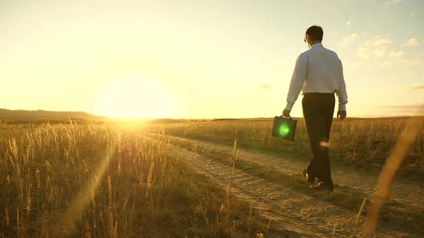 Επιχειρηματίας πηγαίνει σε έναν επαρχιακό δρόμο με έναν χαρτοφύλακα στο χέρι του. Ο επιχειρηματίας δουλεύει σε μια αγροτική περιοχή. ένας αγρότης ελέγχει τη γη το βράδυ στο ηλιοβασίλεμα. αγροτική επιχειρηματική ιδέα. — Φωτογραφία Αρχείου