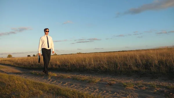 businessman in sunglasses goes down country road with a briefcase in his hand. agricultural business concept. The entrepreneur works in a rural area. farmer inspects the land at sunset. Slow motion