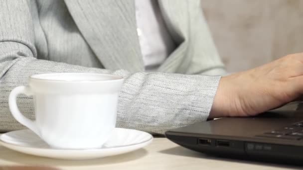 Weiße Kaffeetasse steht auf Schreibtisch. junge Geschäftsfrau, die am Computer arbeitet. Nahaufnahme. Mädchen sitzt im Bürostuhl und tippt auf Laptop. — Stockvideo