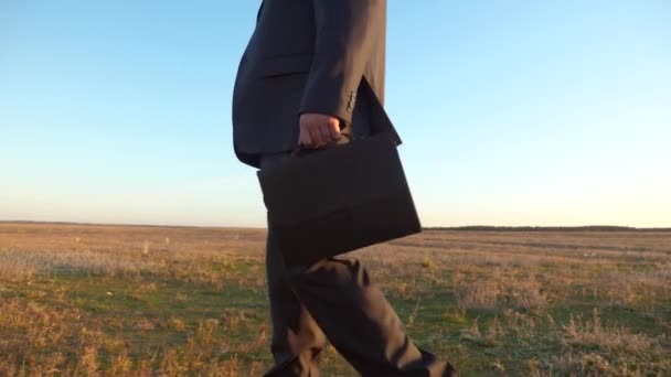 Zakenman in een pak doorloopt park in de stralen van de zonsondergang. Akkerbouwer wandelingen over het veld in de zomer met een zwarte koffer in zijn hand. Close-up. — Stockvideo