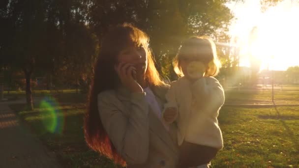 Uma jovem mãe fala em um smartphone e carrega uma filhinha em seus braços. Mãe e filha estão andando no parque ao pôr-do-sol. família férias mãe e bebê . — Vídeo de Stock