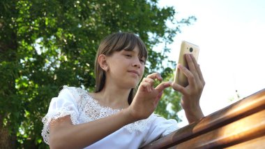 Jestleri ekranın üzerinde yapma Botanik Bahçesi, bin yıllık genç kadında. Bir akıllı telefon kullanan bir genç kız güzel bir yeşil parkta bir bankta bir mektup yazıyor.