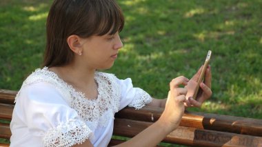 Jestleri ekranın üzerinde yapma Botanik Bahçesi, bin yıllık genç kadında. Bir akıllı telefon kullanan bir genç kız güzel bir yeşil parkta bir bankta bir mektup yazıyor.