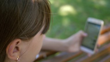 Bir akıllı telefon kullanan bir genç kız, parkta bir bankta bir çevrimiçi mağaza sayfalar arasında saygısız. Bin yıllık kadında genç jestleri ekranın üzerinde yapma Botanik Bahçesi,.