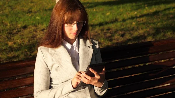 Mulher bonita empresária com óculos verifica e-mail no tablet no parque em um banco — Fotografia de Stock