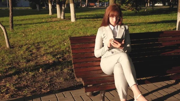female businessman is wearing glasses and is working with tablet and is checking email in summer park on a bench.